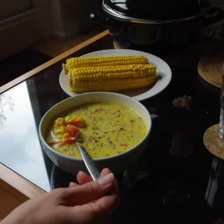 A bowl of corn soup with cobs of corn on a plate behind it