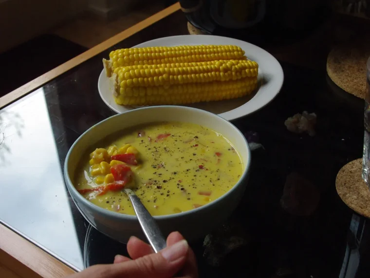A bowl of corn soup with cobs of corn on a plate behind it